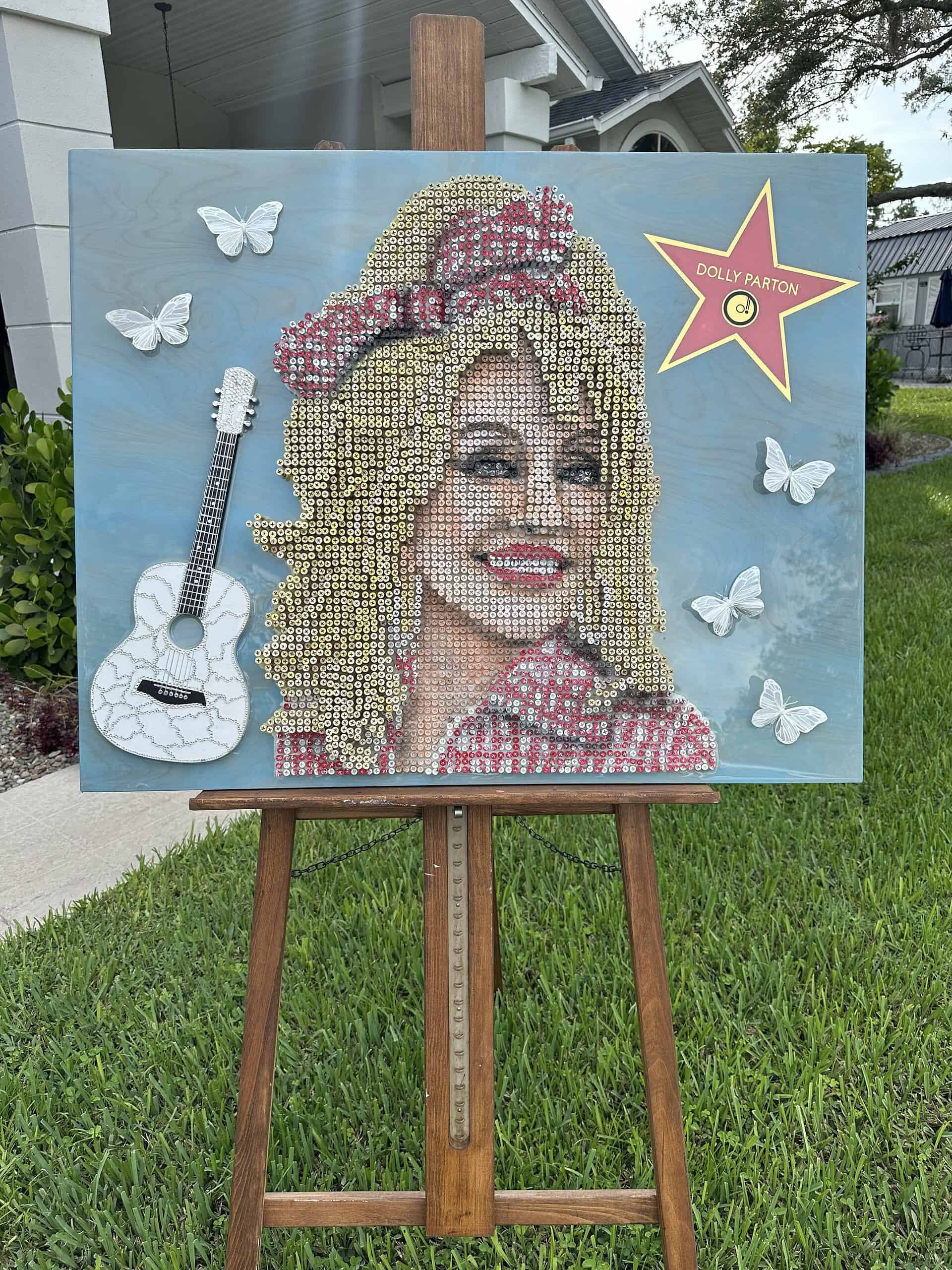 Dolly Parton screw-art piece. Made from over 3,000 screws, 3D butterflies, a miniature replica of her favorite guitar adorned with Swarovski crystals, a Hollywood star, on a wooden blue stained resin coated background! It measures 40” wide x 32” tall. 